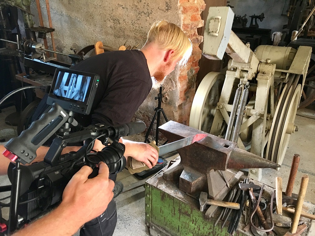 Mann schmiedet mit einem Federhammer und einem Amboss ein Stück Metall. Er wird dabei gefilmt.